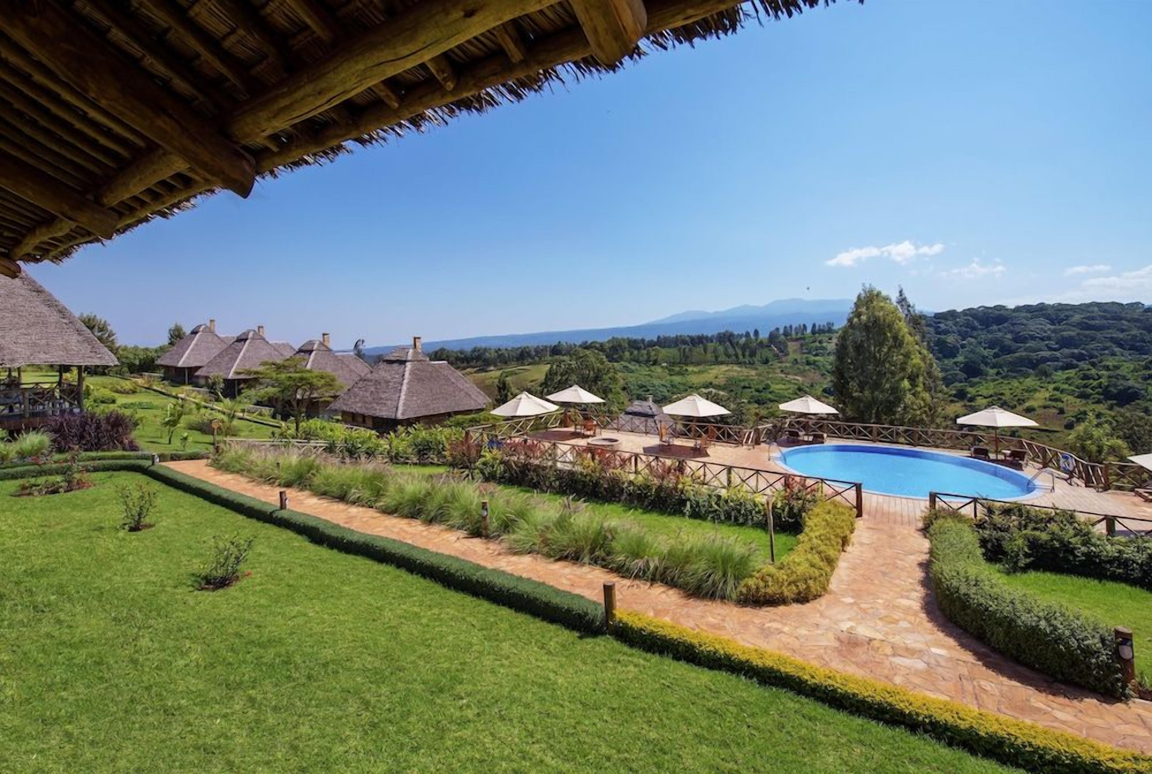 A pool area with a clear view of the surrounding landscape, including a thatched-roof building and a well-maintained lawn.