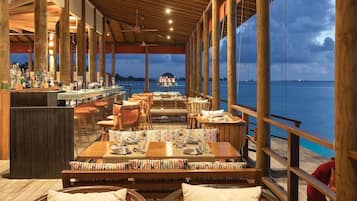 An indoor dining area with wooden furniture, a bar, and large windows overlooking a body of water.