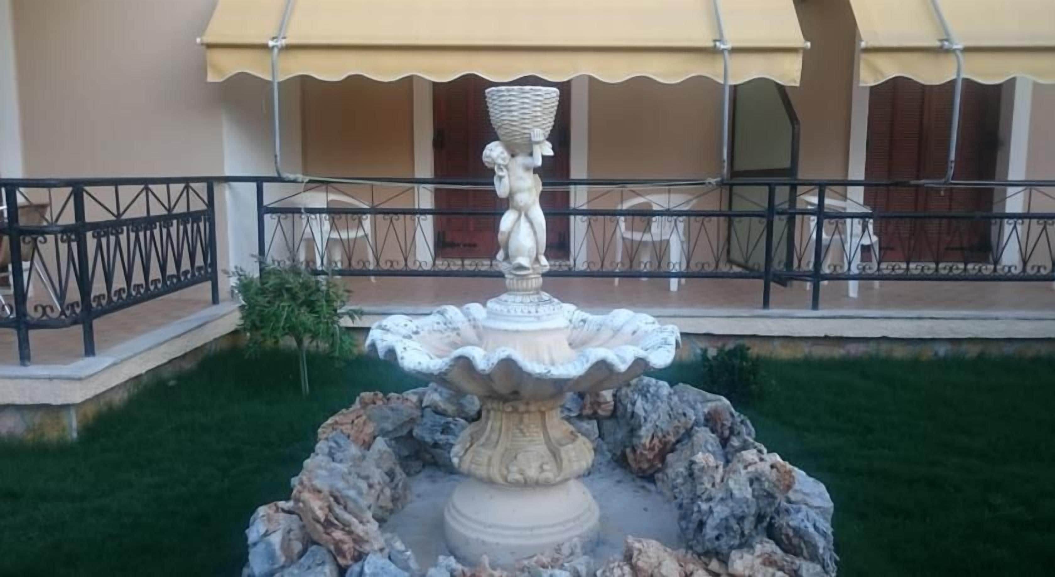 Una fuente con una estatua en el centro, rodeada de rocas y césped, con un edificio al fondo.