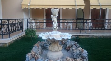 Uma fonte com uma estátua no centro, cercada por pedras e grama, com um edifício ao fundo.
