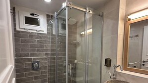 Un bany amb plat de dutxa envoltat de vidre, un vàter blanc, un lavabo amb una banya de pedra i un mirall amb marc de fusta.