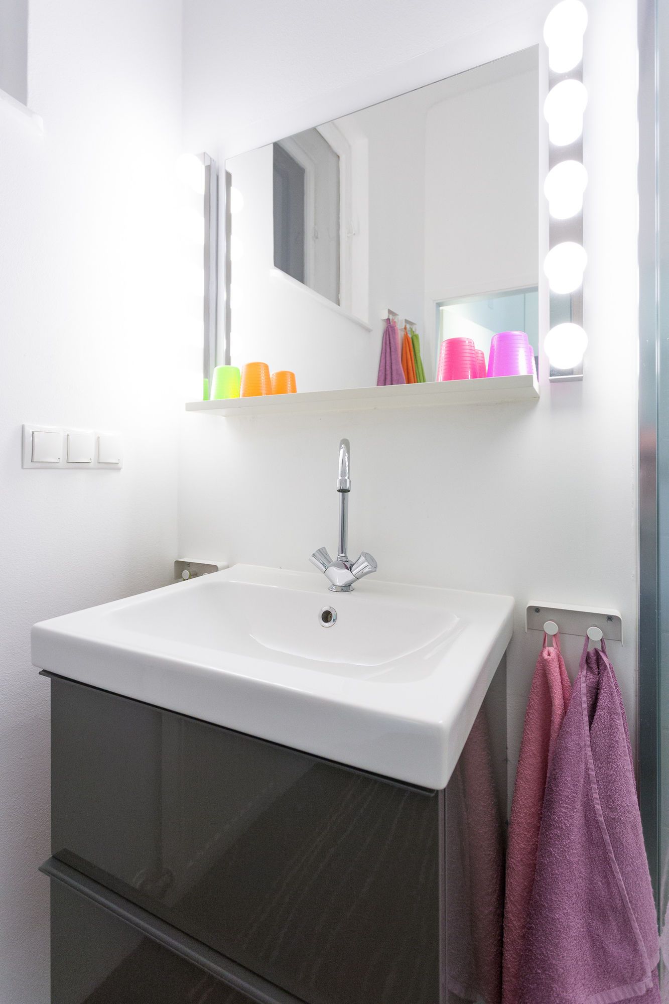 Un baño con un lavamanos blanco, un espejo con luces y tazas de colores en una estantería.
