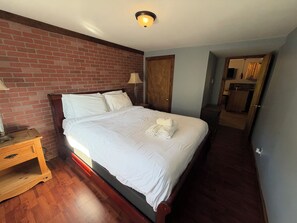 A hotel room with a brick wall, a wooden bed, a nightstand, a lamp, and a bathroom visible through an open door.
