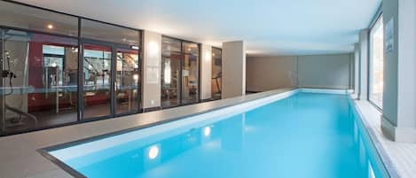 Piscina interior com paredes de vidro transparente, piso revestido e área de exercícios com equipamentos visíveis.