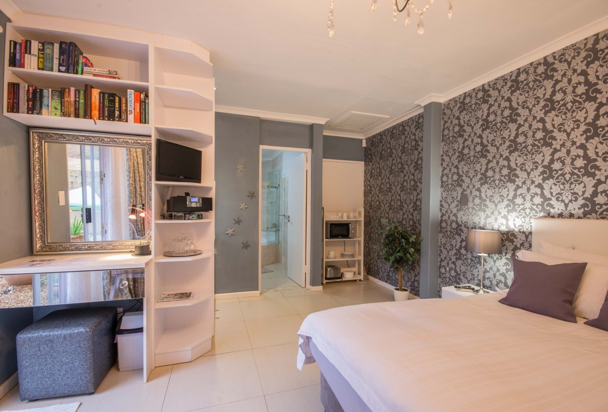 A bedroom with a large bed, a vanity with a mirror, a bookshelf, a television, and a bathroom visible through an open door.