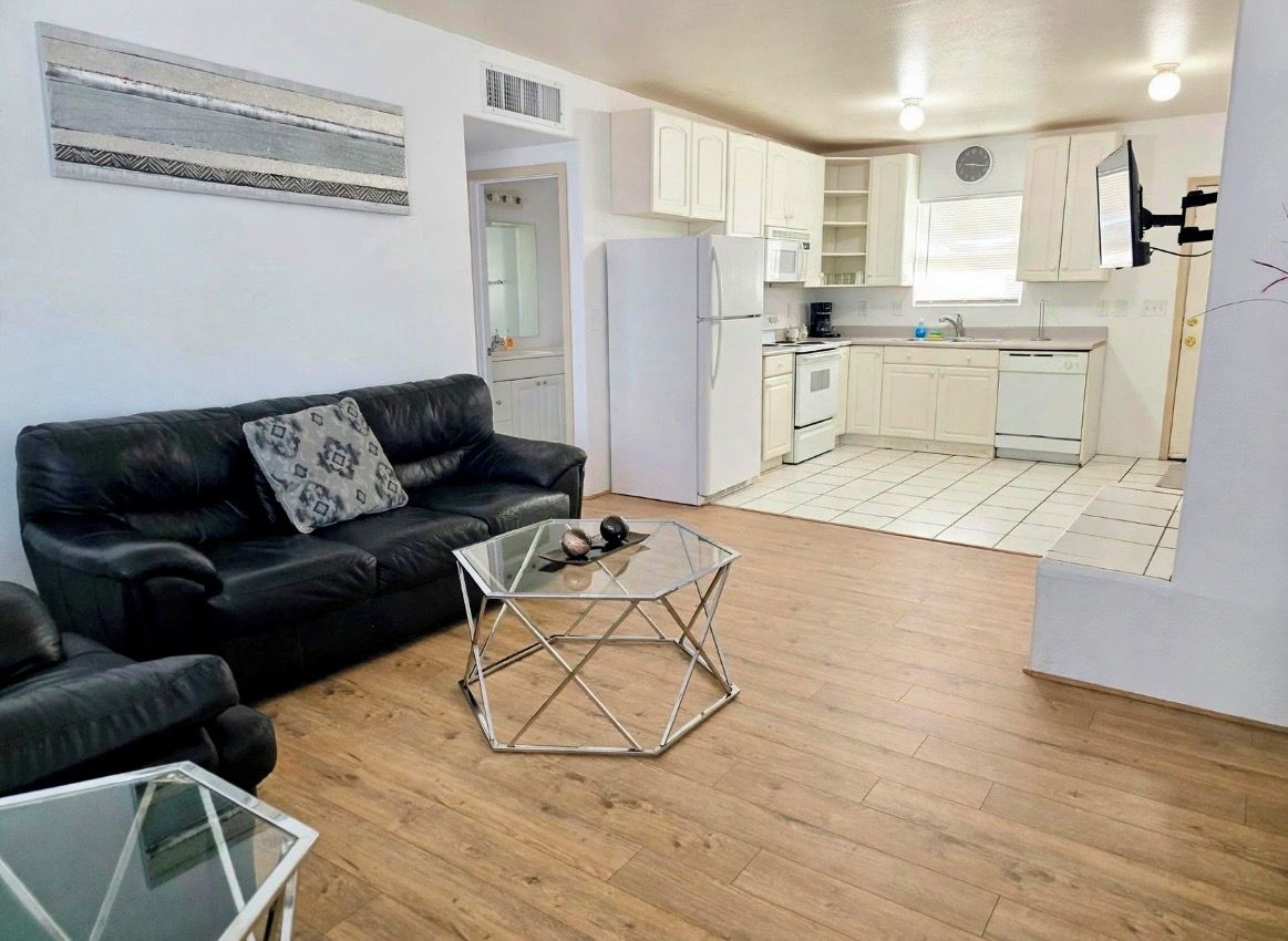 Un espacio habitable compacto con sofá, una mesita de centro de vidrio y una zona de cocina con armarios y electrodomésticos.