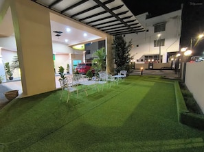 Un espacio exterior con césped artificial, sillas de metal blanco y un coche rojo estacionado dentro de un edificio.
