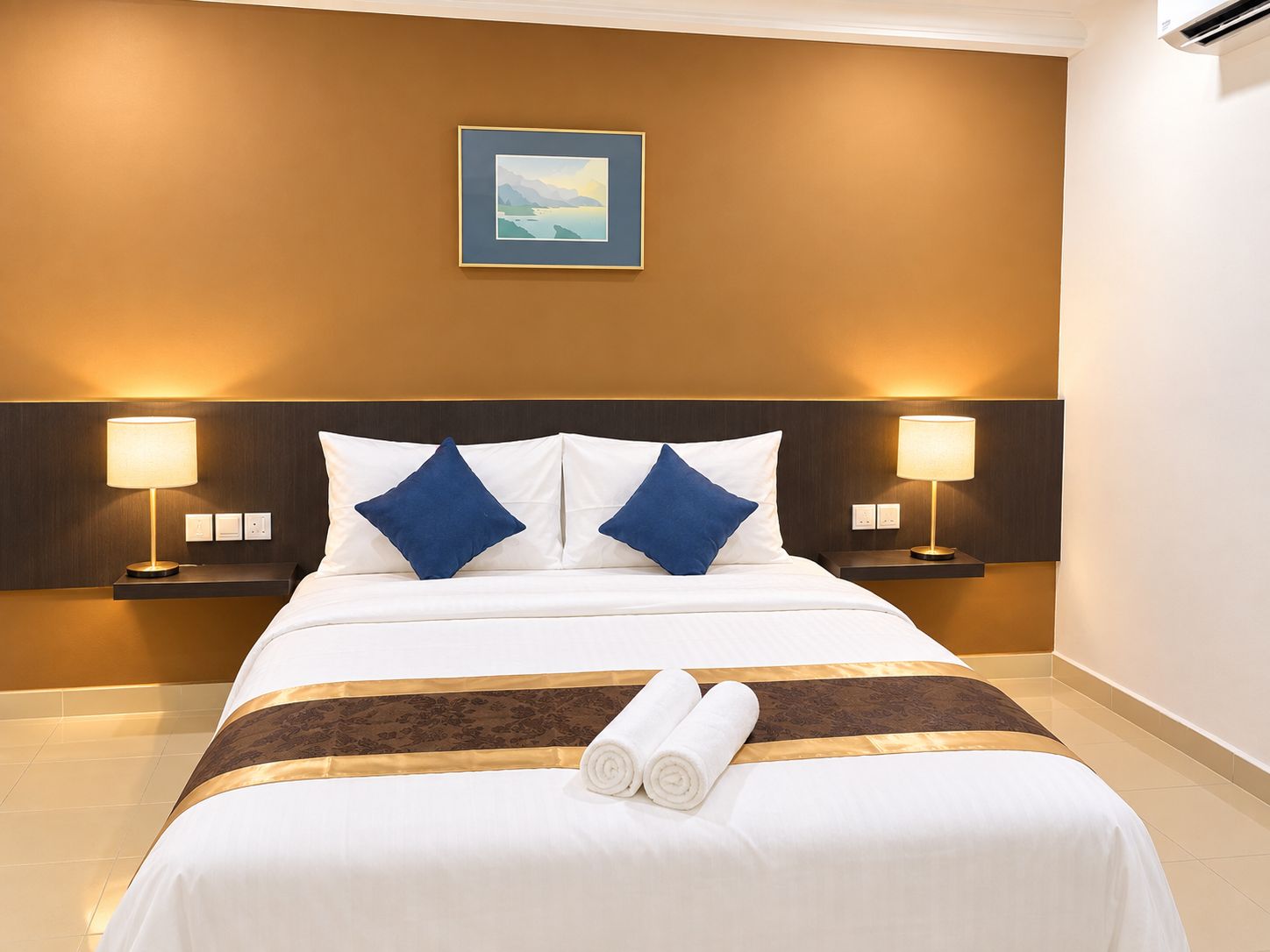 A hotel room with a bed, two bedside lamps, a framed picture on the wall, and rolled towels on the bed.