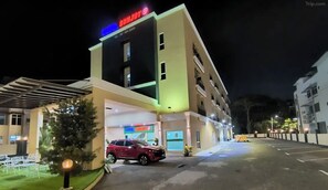Un hotel con un letrero rojo, una zona de estacionamiento y la entrada al edificio.