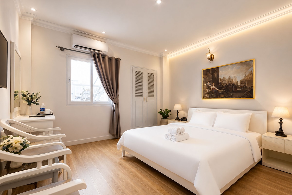 A hotel room with a large bed, bedside tables, a desk with a chair, a window with curtains, an air conditioning unit, and a painting on the wall.