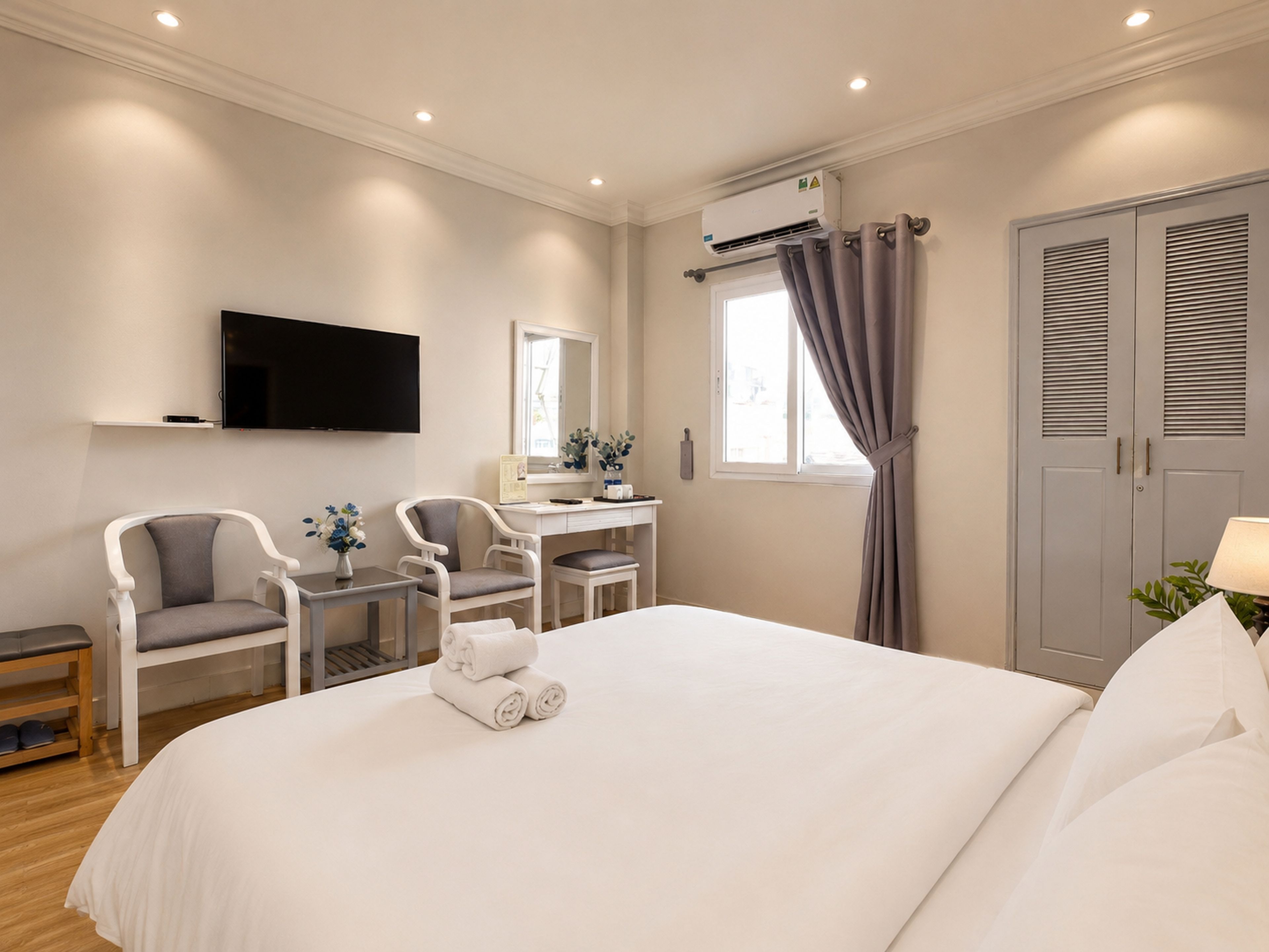 A hotel room with a bed, a desk, two chairs, a television, an air conditioning unit, a window with curtains, and a wardrobe.