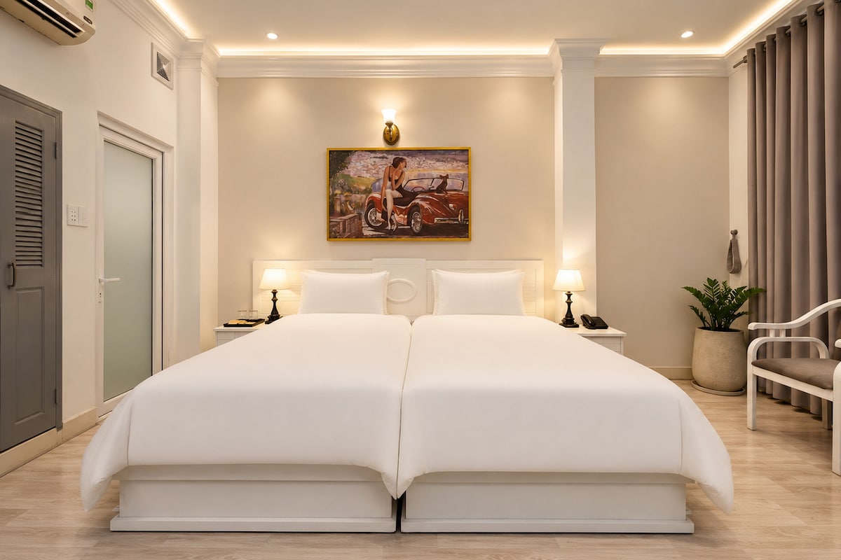 A hotel room with a large bed, two bedside tables, a painting on the wall, a potted plant, and a window with curtains.