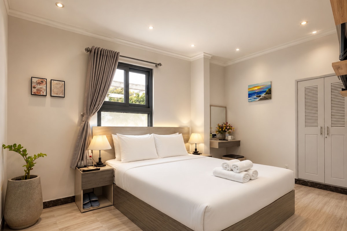 A hotel room with a large bed, bedside lamps, a nightstand, a potted plant, and a window with curtains.