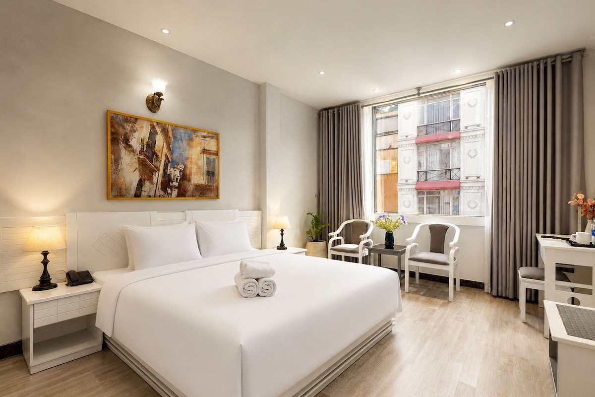 A hotel room with a large bed, a desk, two chairs, a painting on the wall, and a window with curtains.