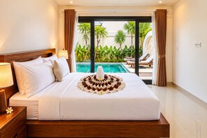 A hotel room with a bed, a nightstand, a lamp, a decorative towel arrangement, and a view of a pool and greenery outside. - Viondra Villas Canggu (Badung)