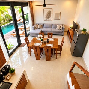 A modern living room with a grey sofa, a dining table set for four, and a kitchen area with a refrigerator. - Viondra Villas Canggu (Badung)