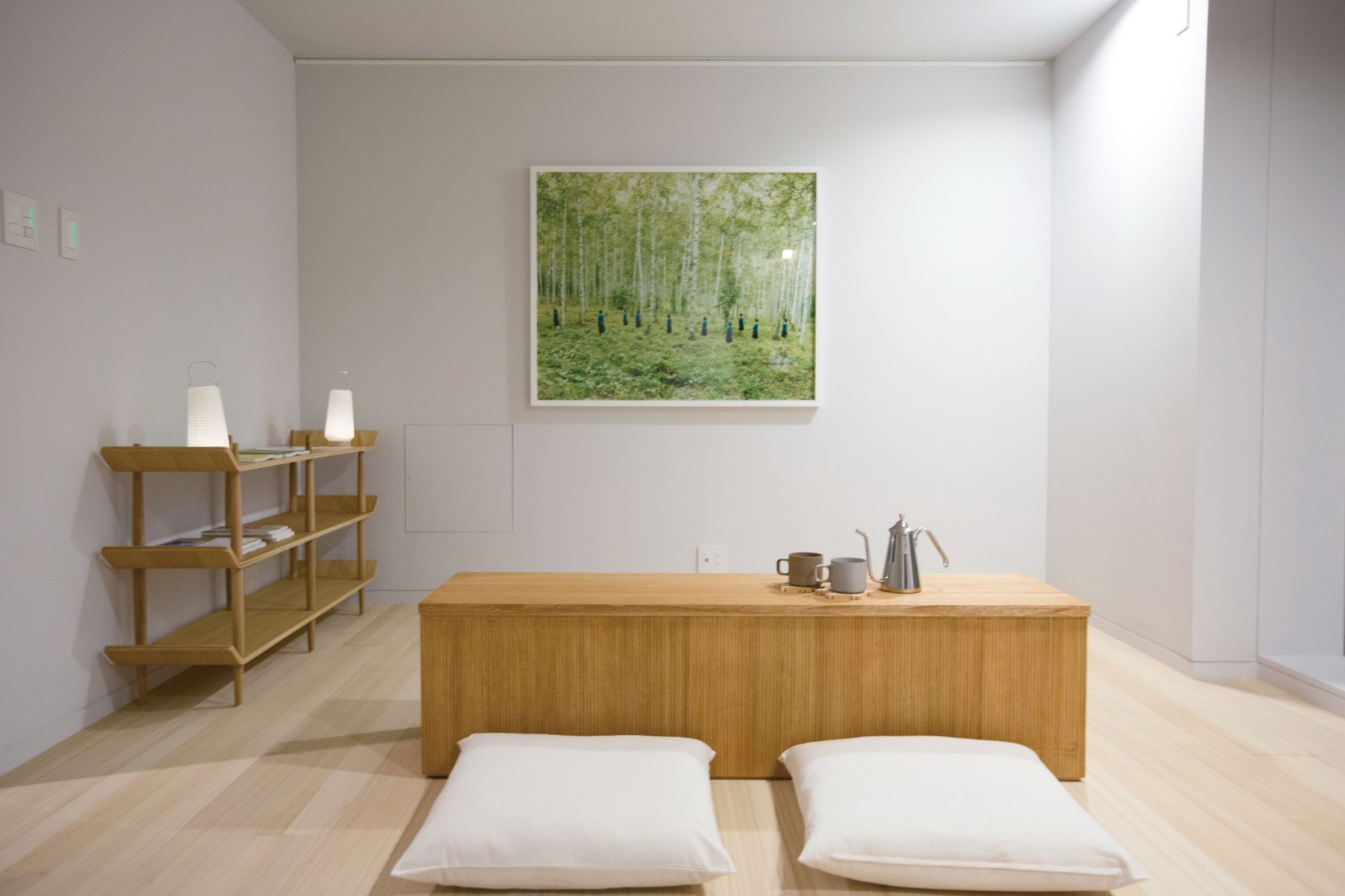 A minimalist room with a wooden bench, two white cushions, a shelf with books and a lamp, and a painting of a forest.
