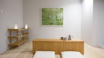A minimalist room with a wooden bench, two white cushions, a shelf with books and a lamp, and a painting of a forest.