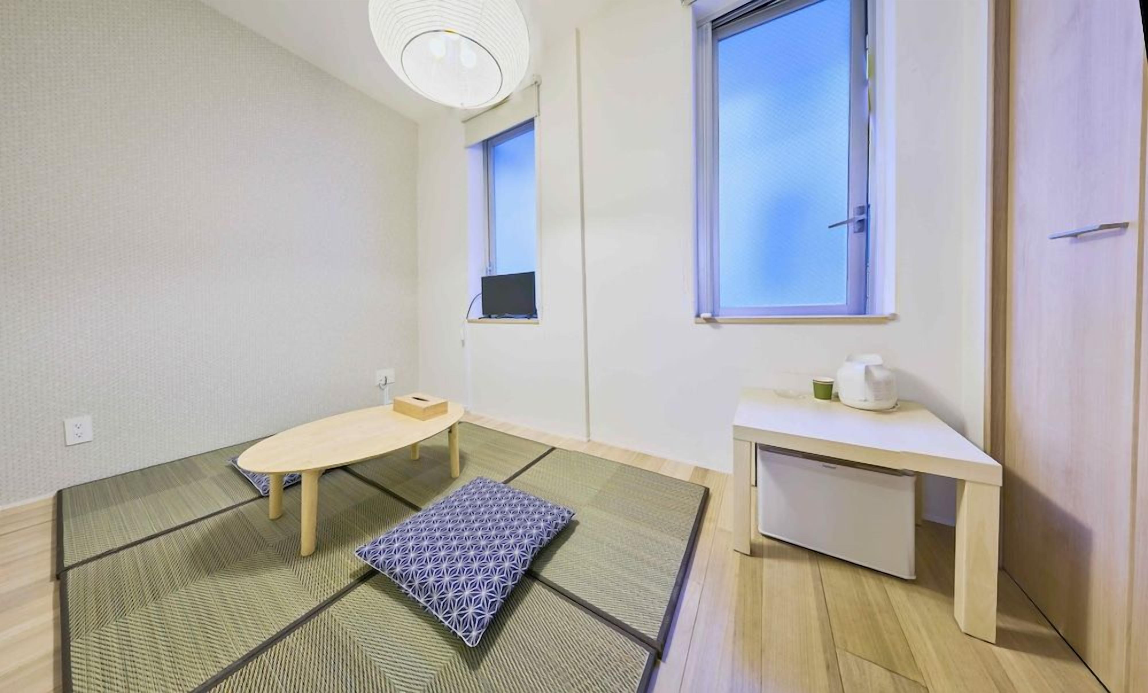 A room with tatami mats, a low wooden table, a small bench, a white desk with a cup, and a window with blinds.