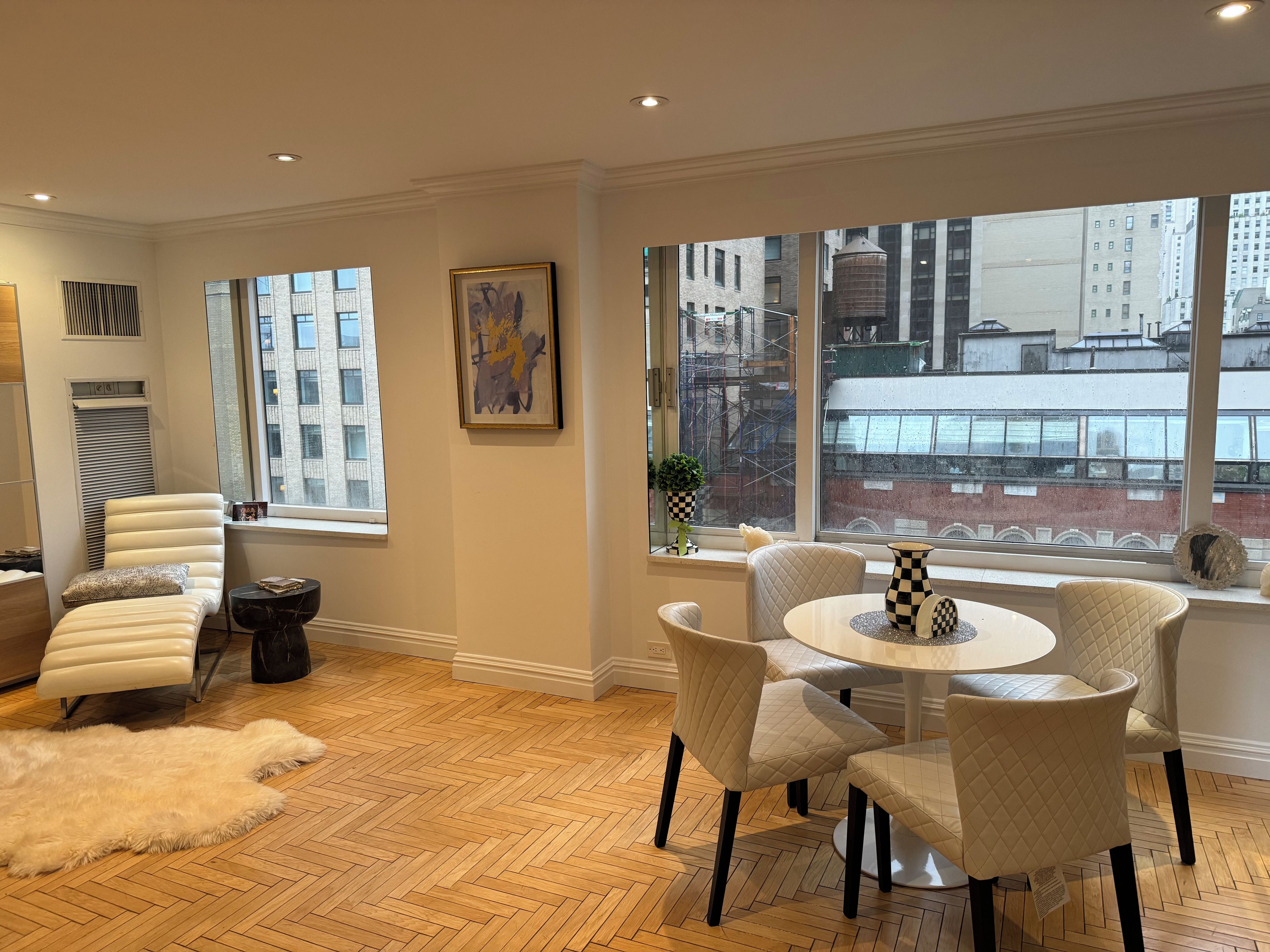 A modern living room with a large window, a white leather sofa, a round table with chairs, and a herringbone wooden floor.