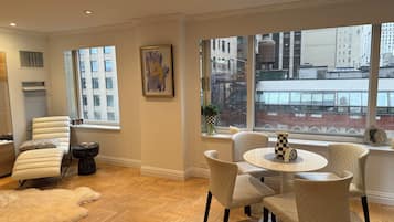 A modern living room with a large window, a white leather sofa, a round table with chairs, and a herringbone wooden floor.
