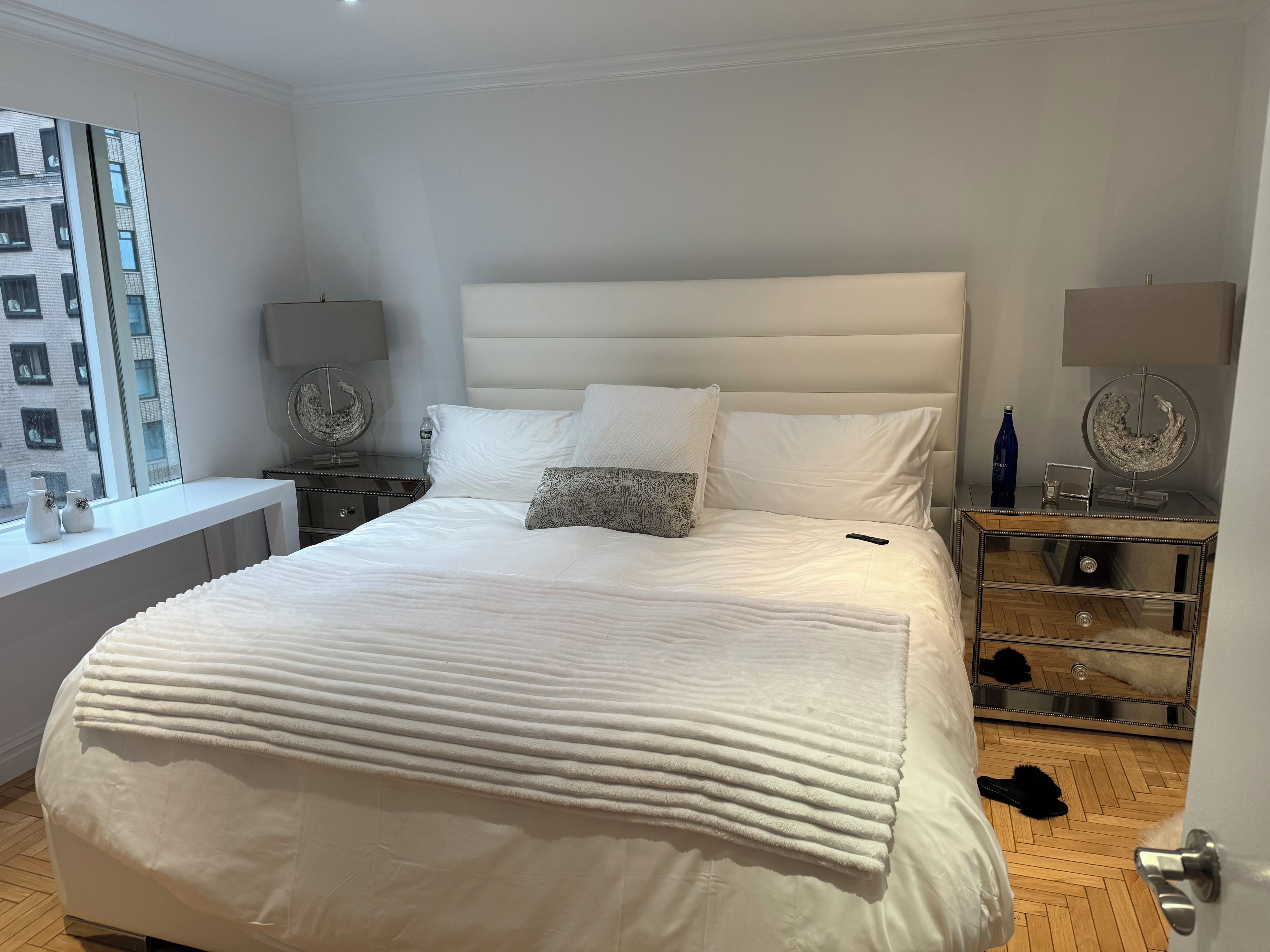 A bedroom with a large bed, a bedside table with a lamp, a mirror, and a view of a building through a window.