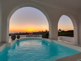 Piscina con vista al atardecer, rodeada de columnas blancas y un área de descanso con cojines y una cesta.