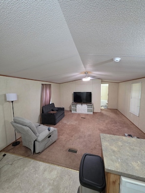 A compact living space with a ceiling fan, a flat-screen TV, a sofa, a massage chair, and a small kitchenette area.