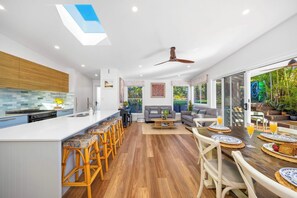 A modern kitchen with a central island, bar stools, and a dining area with a wooden table and chairs. - Green Oasis - Renovated family home (Port Macquarie)