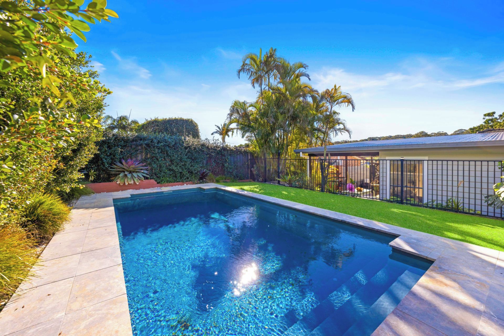 A swimming pool with a tiled deck, surrounded by lush greenery and a fenced area.