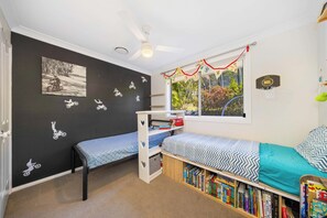 Ein Kinderzimmer mit zwei Etagenbetten, einem Bücherregal, einem Deckenventilator und einem Fenster mit Blick auf Grünflächen.