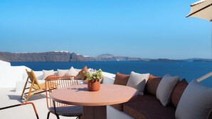A round table with chairs and a sofa, a potted plant, and a view of the sea and distant land. - Santa Irini Villa (Santorini)