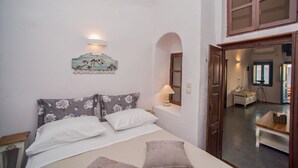 A bedroom with a bed, a nightstand, a lamp, a decorative wall piece, and a view of the adjacent room. - Santa Irini Villa (Santorini)