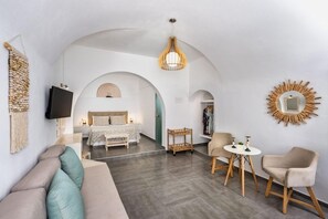 Habitació d'hotel moderna amb sofà, llit amb capçalera, televisió, taula de dinar amb cadires i un mirall decoratiu.
