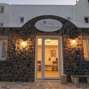 En bygning i sten med en facade, hvor der står 'Villa Thiranthemis' og en dør, der fører ind i et rum med et bord og stole.