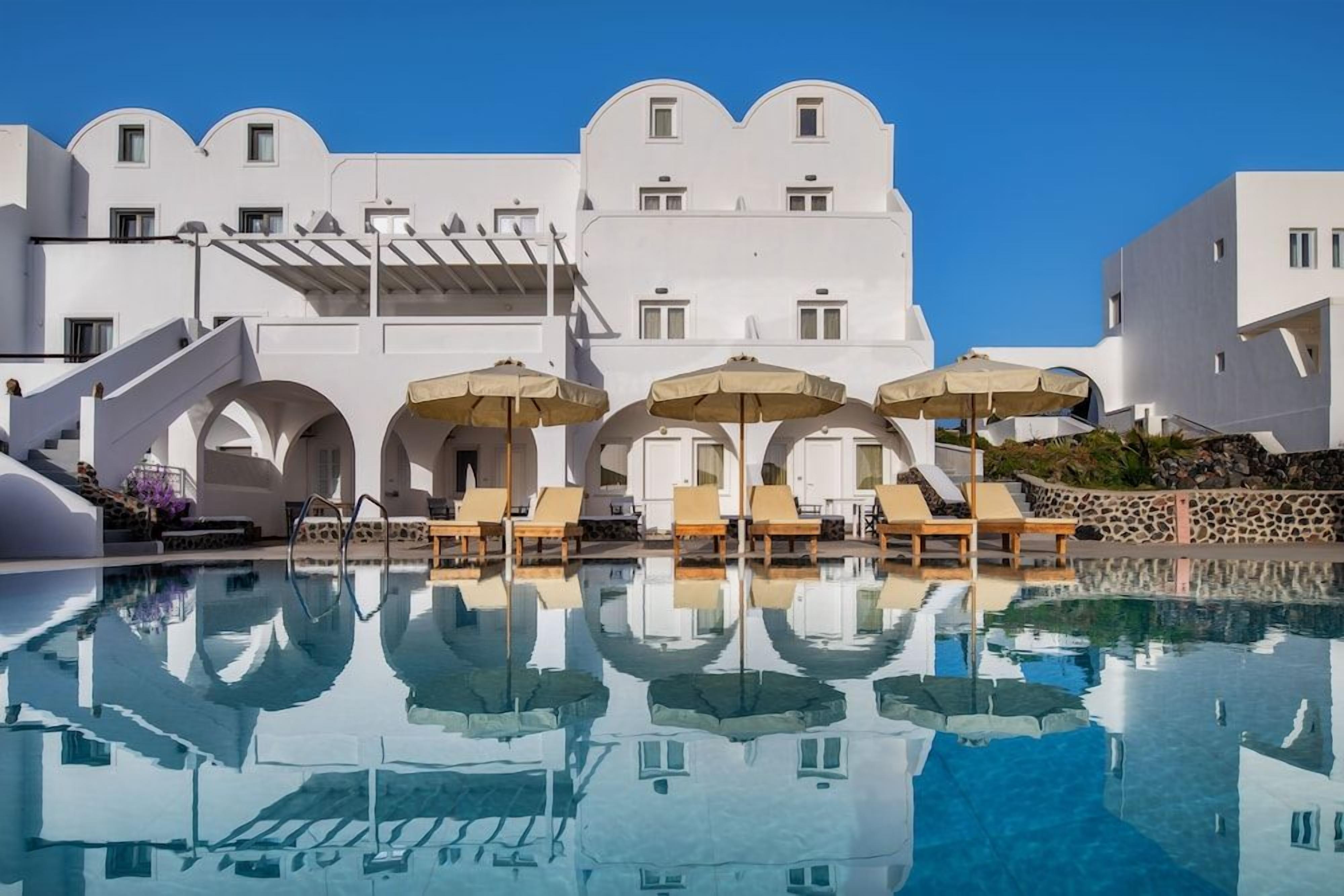 A poolside area with lounge chairs and umbrellas, a clear pool reflecting the building, and a white building with arches and balconies.
