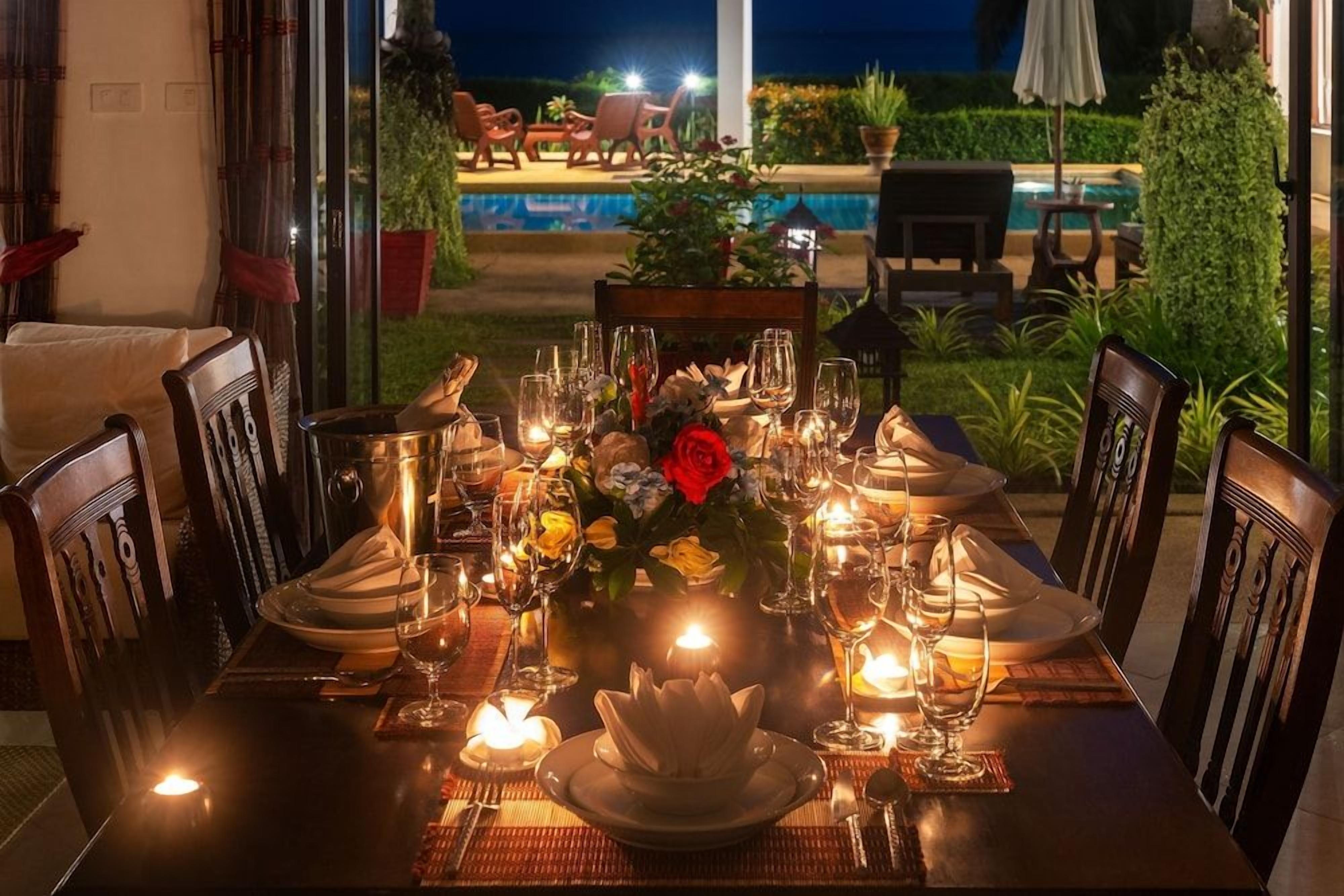 An outdoor dining area with a table set for a meal, surrounded by chairs, with a pool and garden in the background.