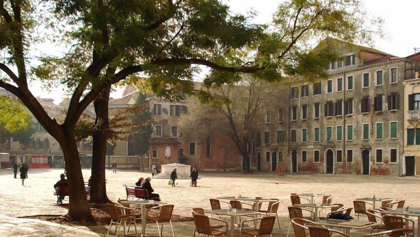 An outdoor seating area with tables and chairs, surrounded by trees and buildings. - Red Rose (Venice)