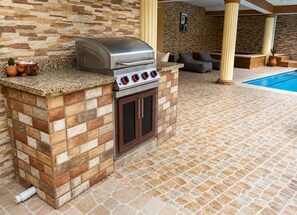 Une cuisine extérieure dotée d’un barbecue et d’un four intégrés, une piscine en arrière-plan et un sol carrelé en pierre.