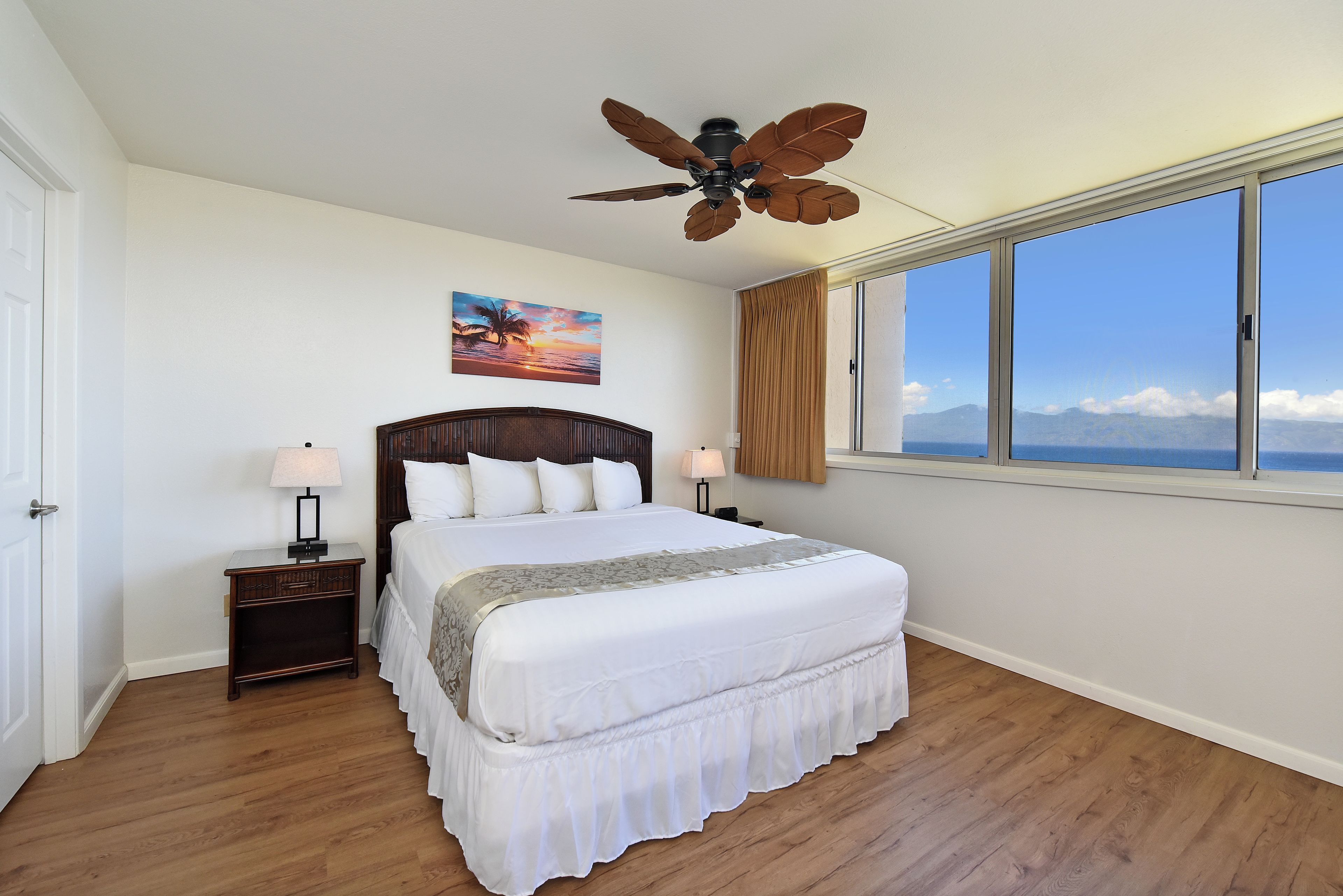 Quarto com cama, mesa de cabeceira, ventilador de teto, janela com vista e um quadro na parede.