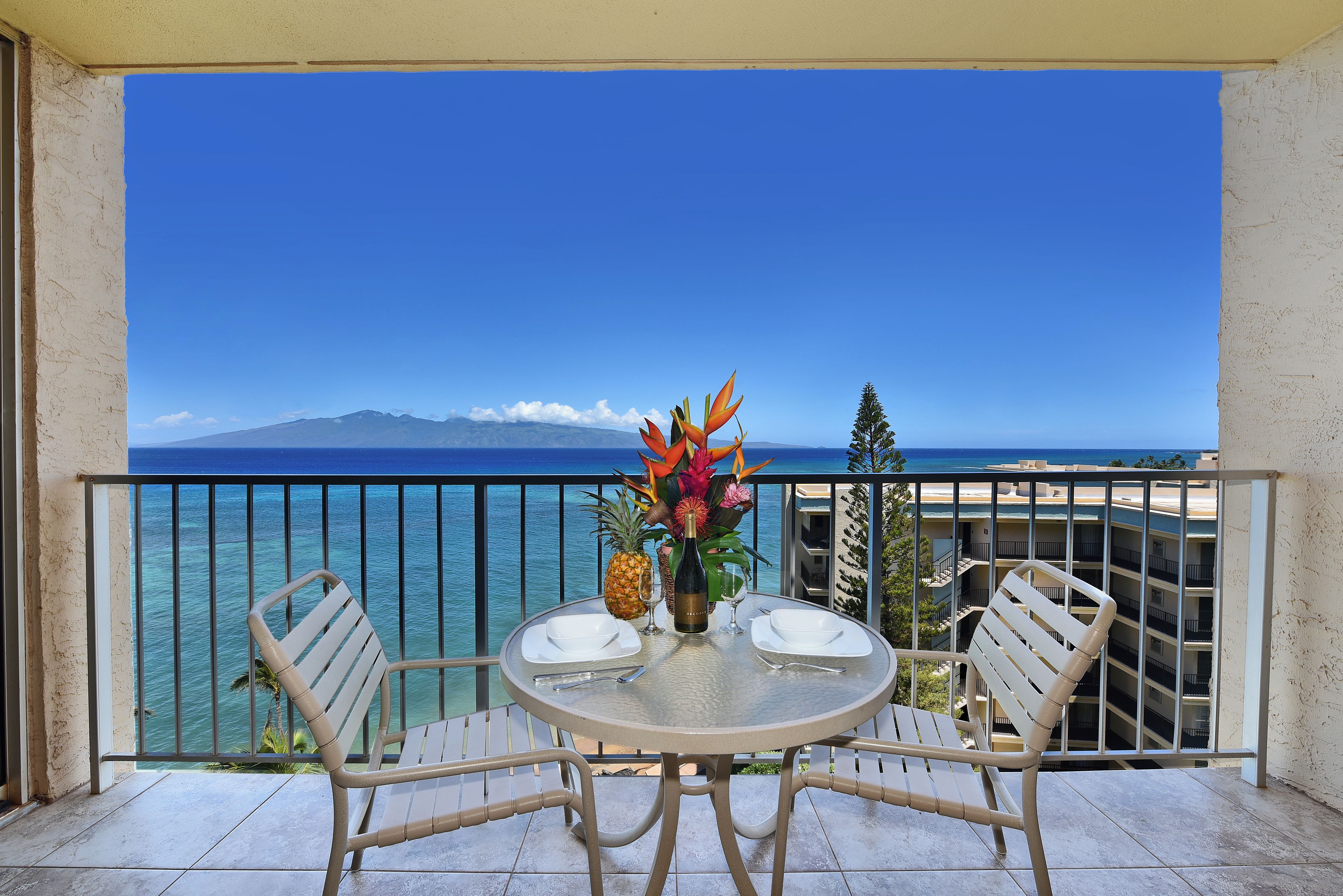 A balcony with a table set for two, overlooking a clear blue sea and a distant mountain range.