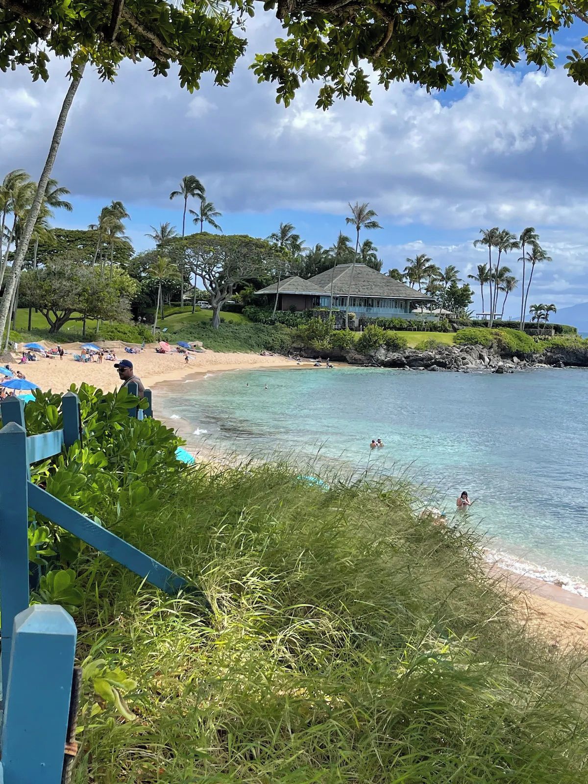 A beach with clear turquoise water, a wooden fence, and a house with a thatched roof in the background.