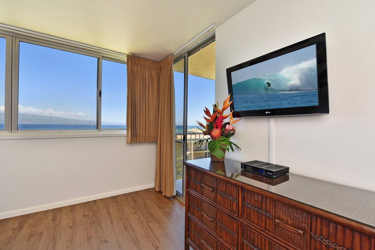A room with a large window, a flat-screen TV, a wooden dresser, and a plant.