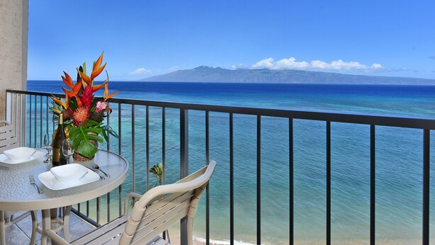 A balcony with a table set for dining, overlooking a clear blue sea and distant mountains. - Oceanfront - Luxury View - 2 Beds (Lahaina)