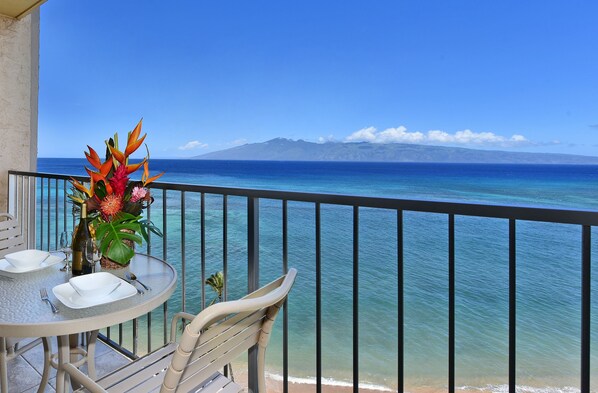 Un balcon avec une table dressée pour le dîner, offrant une vue sur une mer bleue et limpide et sur des montagnes lointaines.