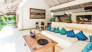 A spacious living area with a wooden coffee table, white sofa with patterned cushions, and a decorative wall piece. - Villa Gembira (Keroboken Kelod)