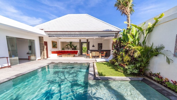 Una casa moderna amb una piscina, un pati amb mobles d'exterior i una gran varietat de plantes tropicals.