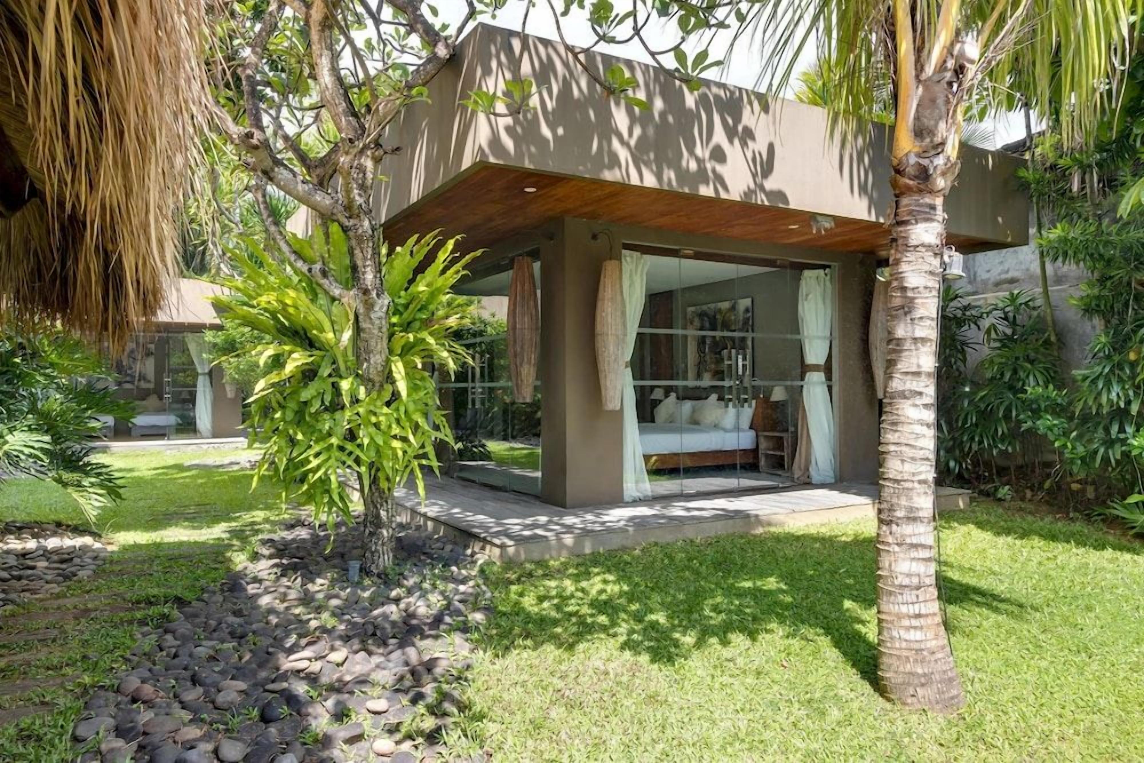 A modern house with a glass-enclosed porch, a bed, and a small garden with palm trees and a stone pathway.
