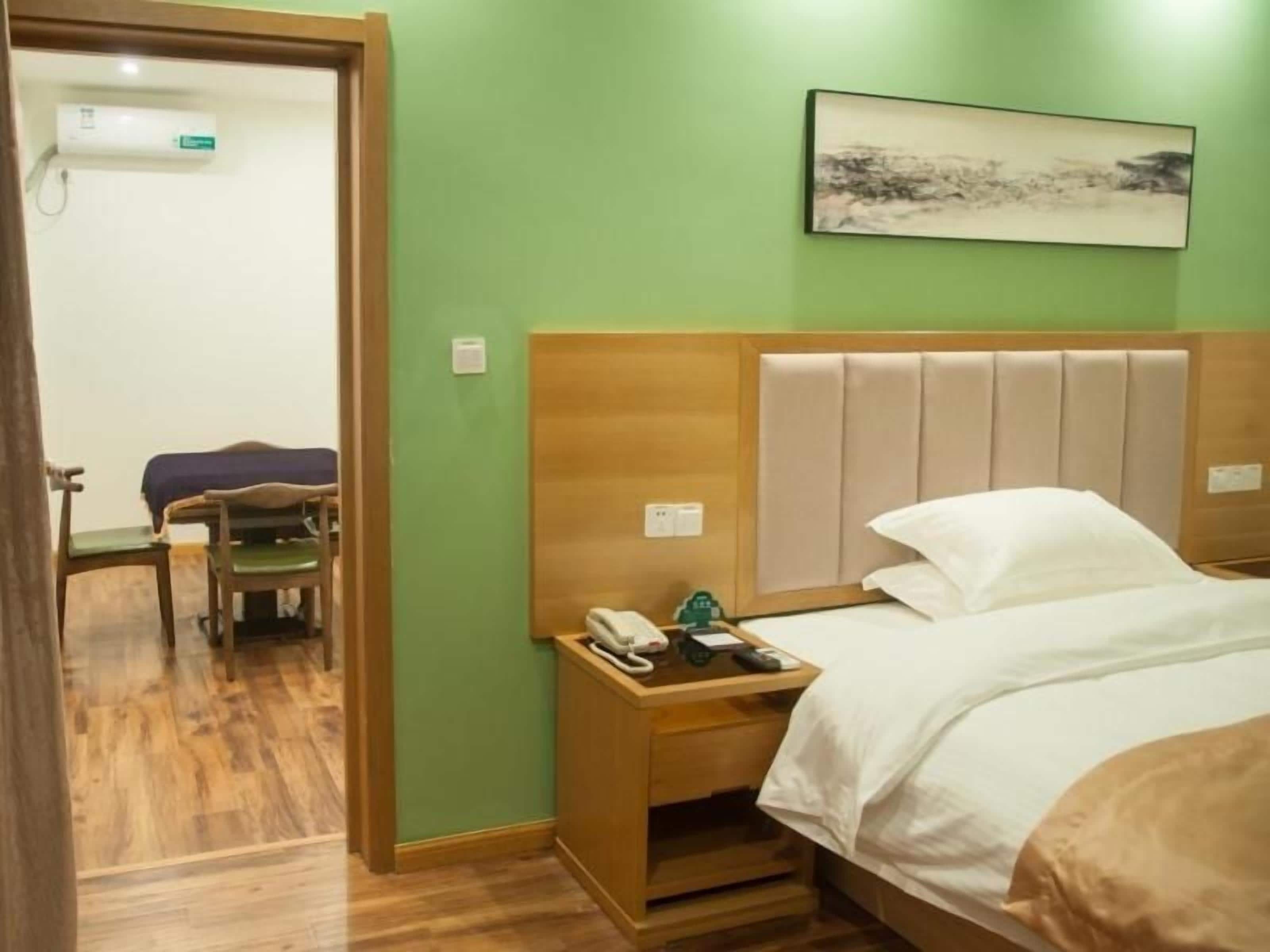 A hotel room with a wooden headboard, a bedside table with a phone and a green object, a bed with white bedding, and a painting on the wall.