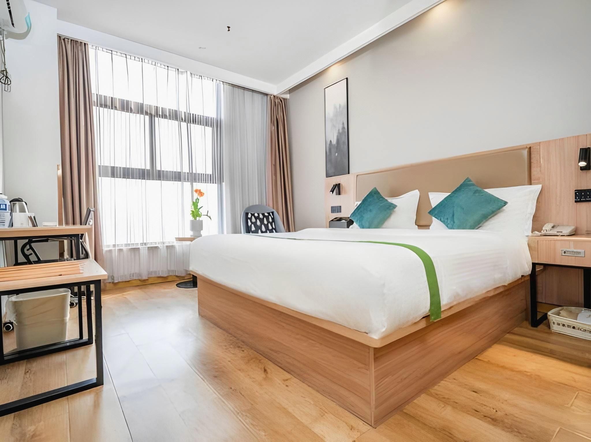 A hotel room with a wooden bed, a desk, a chair, a bedside table, a lamp, and a window with curtains.